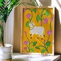 Framed artwork of a cow and flowers on a yellow background, placed on a surface with a plant beside it.