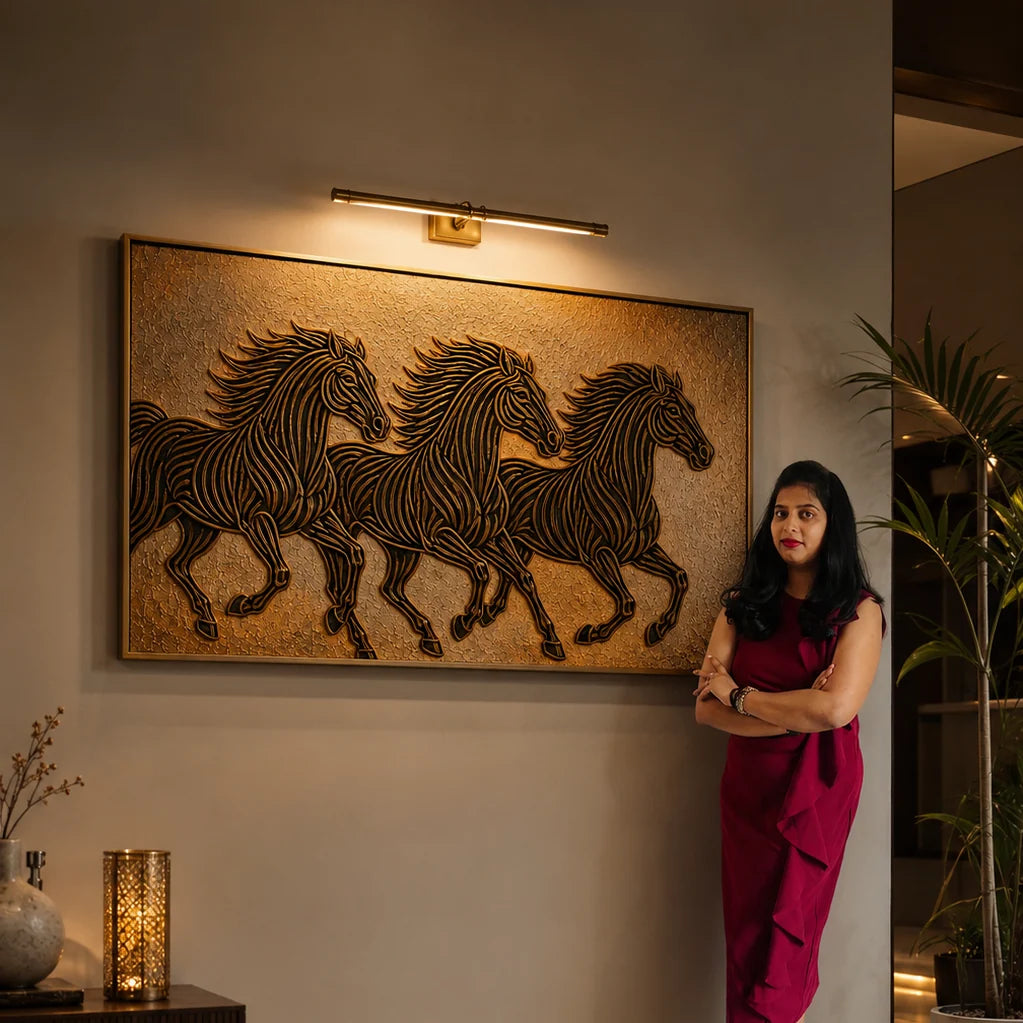 A woman in a burgundy dress stands beside a large framed wall art piece showing three black horses on a textured gold background, mounted on a beige wall under a modern light fixture.